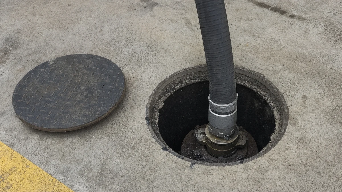 Mangote de descarga preto ligado por engate rápido ao bocal do tanque subterrâneo de um posto, dentro do poço de contenção aberto no piso de concreto, com a tampa de ferro ao lado